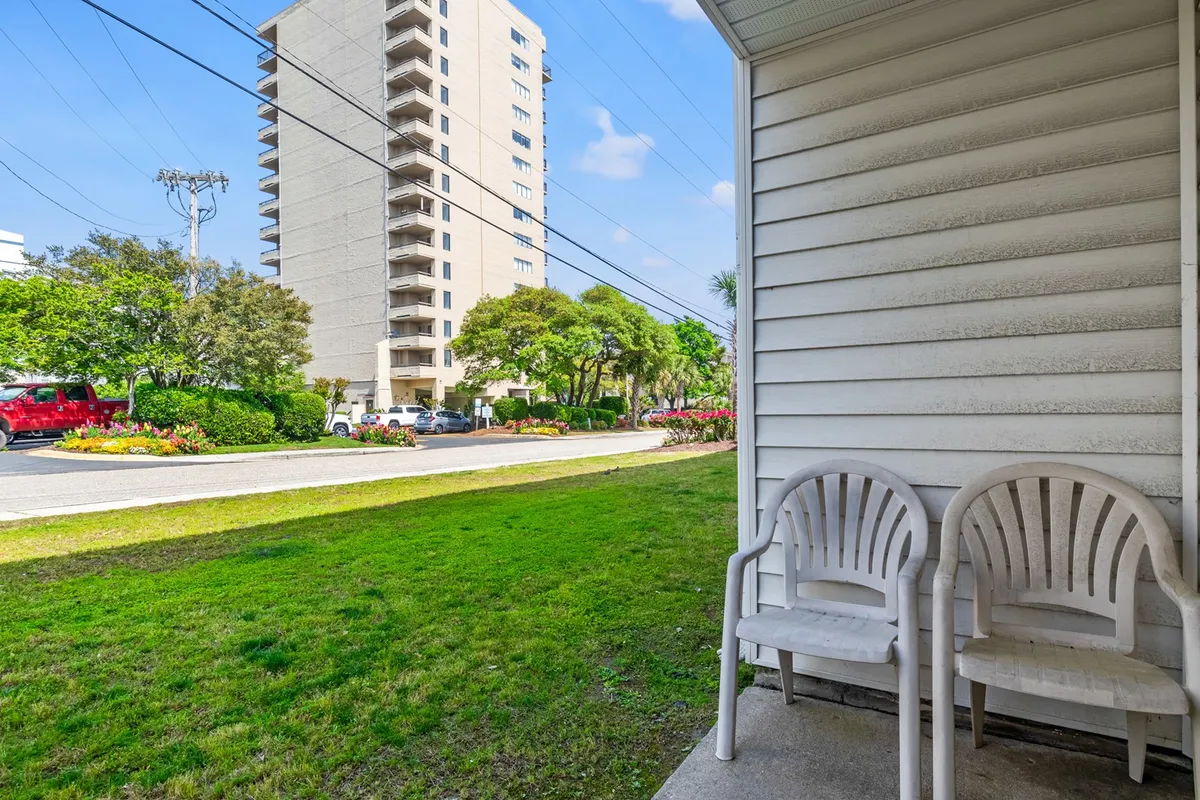 This exterior image features two plastic chairs set on a balcony or porch surrounded by landscape greenery, with the apartment building visible in the background. The scene is tranquil, inviting relaxation, and directly evokes the experience of enjoying a morning coffee with fresh air.
