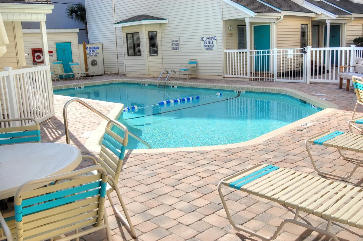 The hero image showcases a sparkling swimming pool surrounded by inviting lounge chairs, a bright white fence, and charming cabana-style condo buildings. The atmosphere is sun-soaked and relaxing, encouraging guests to imagine unwinding by the water with friends or family.