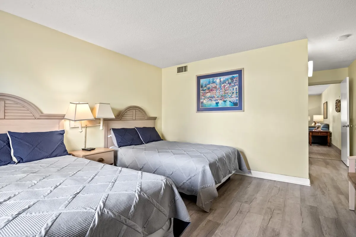 This image features two beds dressed in soft linens, flanked by nightstands with lamps and complemented by framed artwork. The lighting and arrangement create a warm, inviting space ideal for relaxing, socializing, or winding down after a long day.
