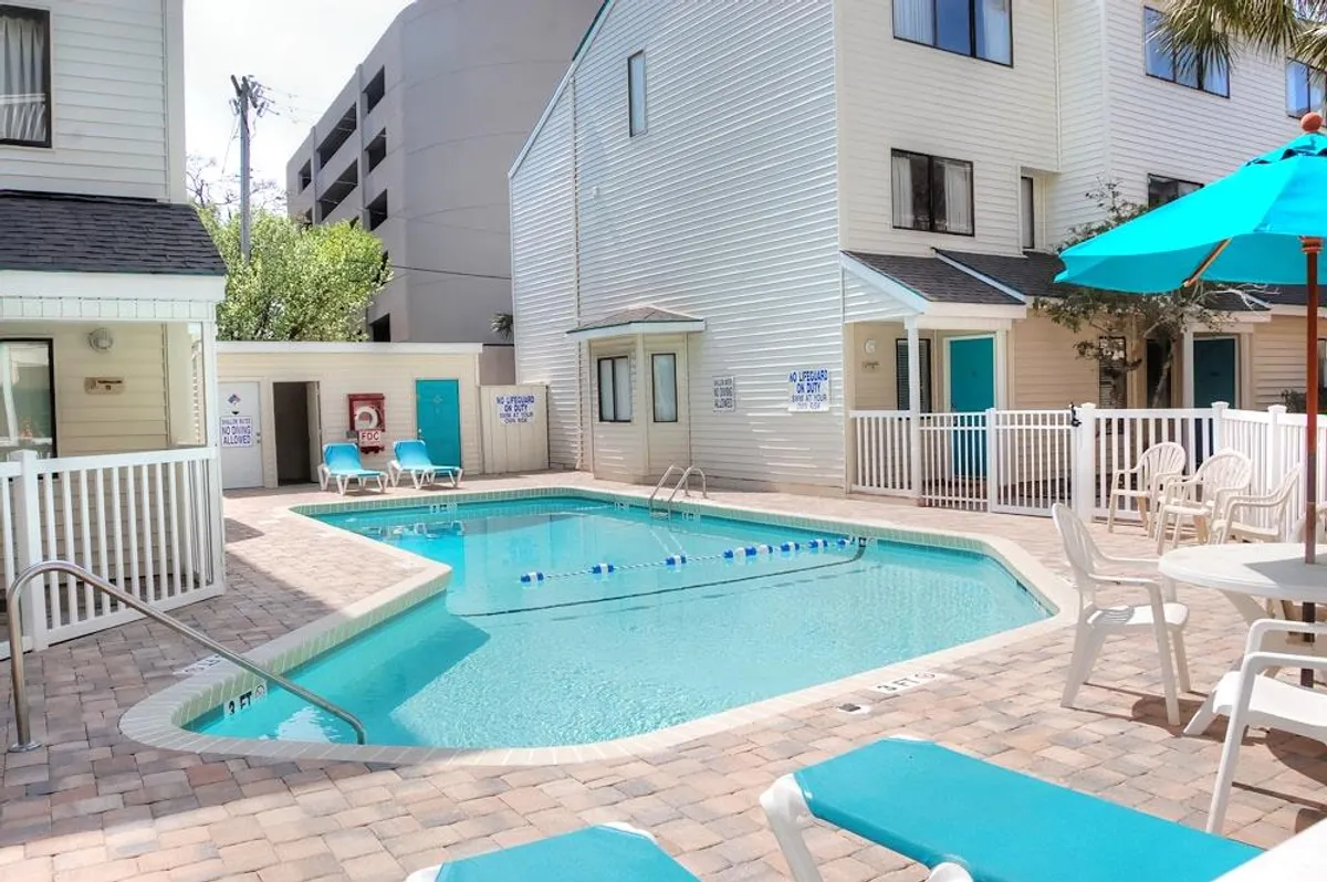 An inviting outdoor swimming pool is surrounded by lounge chairs and umbrellas, with resort buildings visible in the background. The setting feels relaxed and cheerful, offering a glimpse of the on-site amenities that add value to a guest's stay.