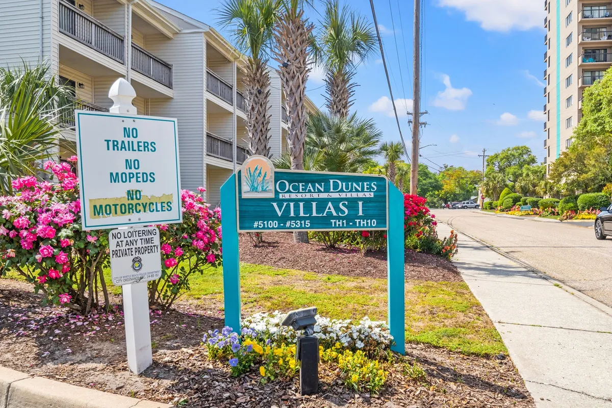 This inviting image features the Ocean Dunes sign set amid vibrant flowers, palm trees, and the building’s distinctive architecture. The scene radiates a welcoming, resort-style atmosphere, suggesting arrival and easy access to both the property and nearby attractions.