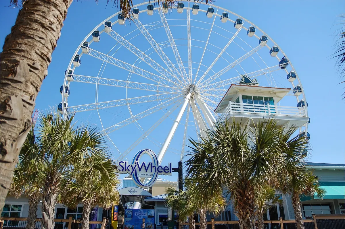 This image captures the iconic SkyWheel in Myrtle Beach, framed by palm trees and a ticket booth at the base. The vibrant scene is lively and full of energy, reflecting one of the area's top attractions included in the Free Fun Program.