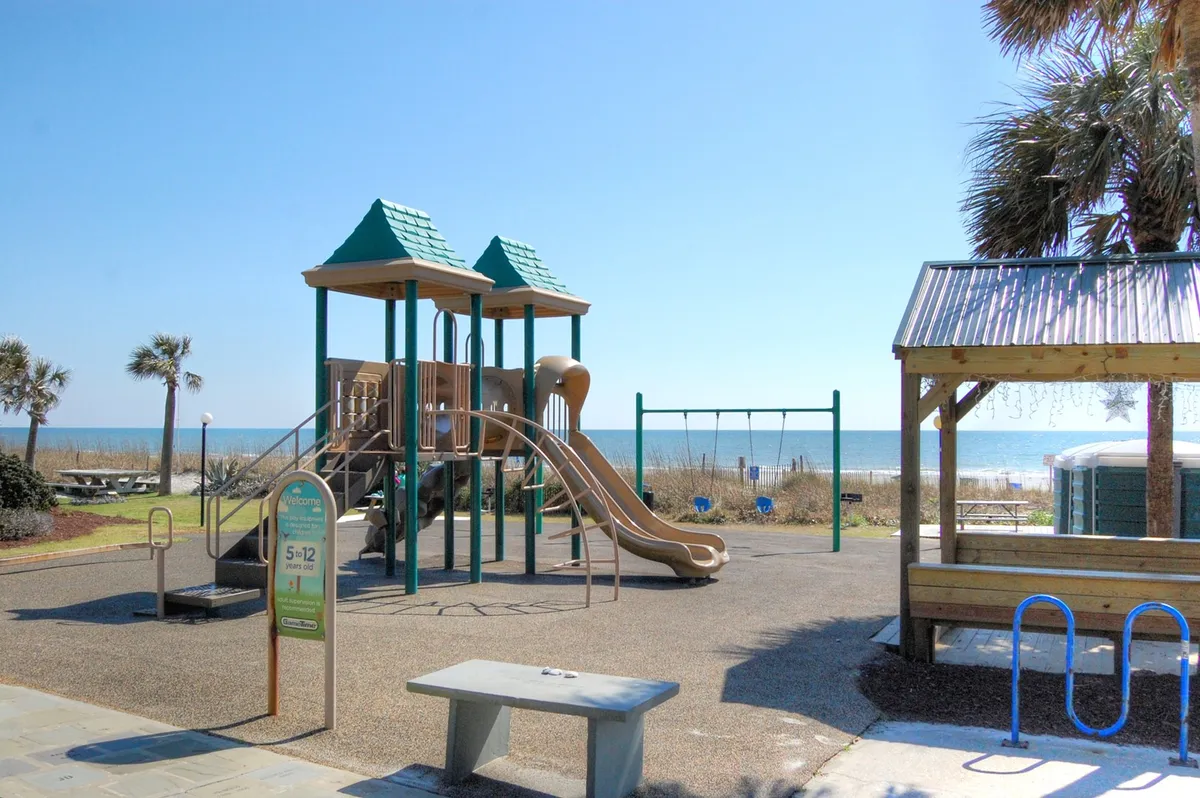 This vibrant exterior image showcases playground equipment, swings, a picnic area, palm trees, and a clear view of the beach. Children can be seen playing while families gather nearby, capturing the essence of outdoor, family-friendly fun close to the shore.