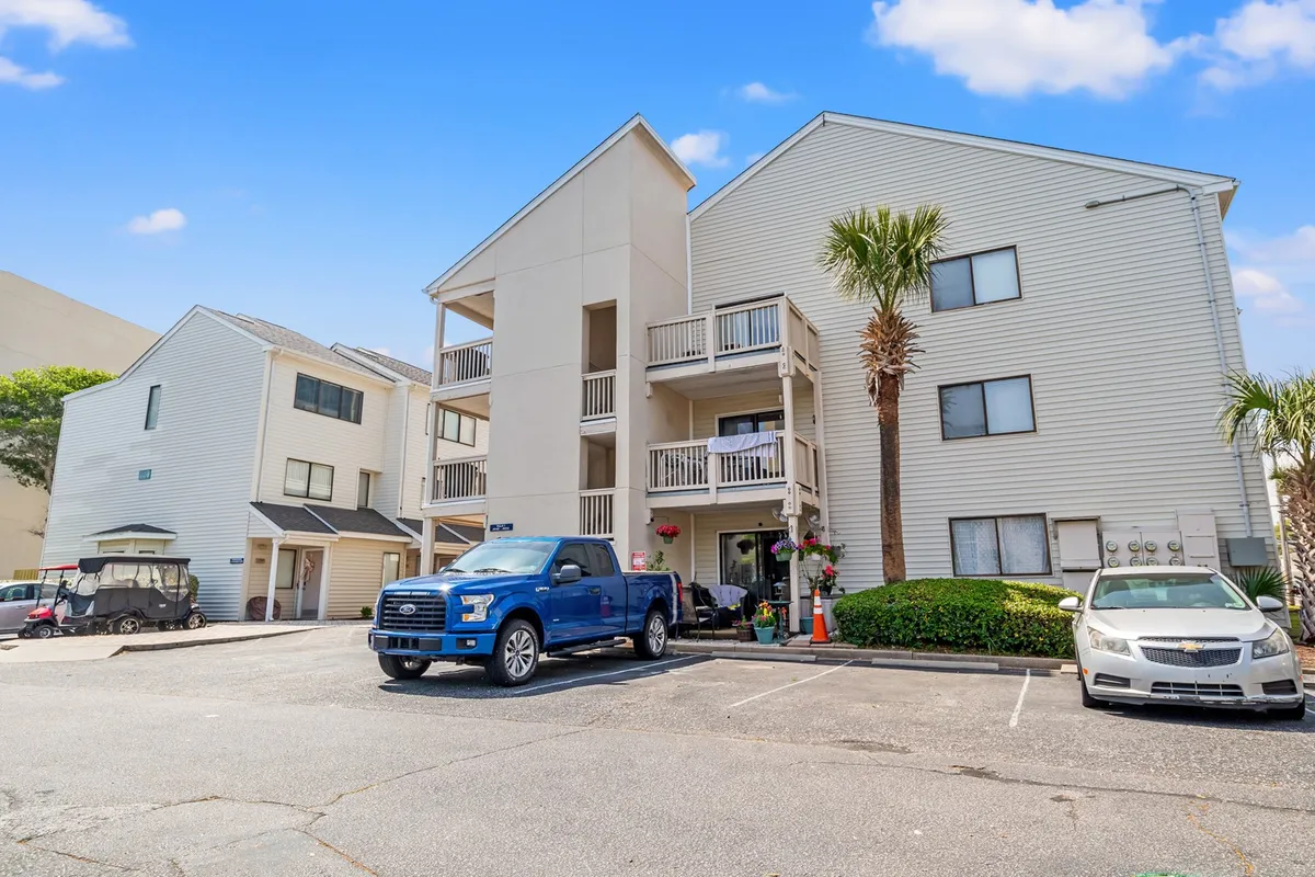 The exterior of the building is highlighted with visible balconies, a blue truck parked below, palm trees, and flower pots. There’s a feeling of friendly community, with residents able to enjoy the outdoors from their own balconies.