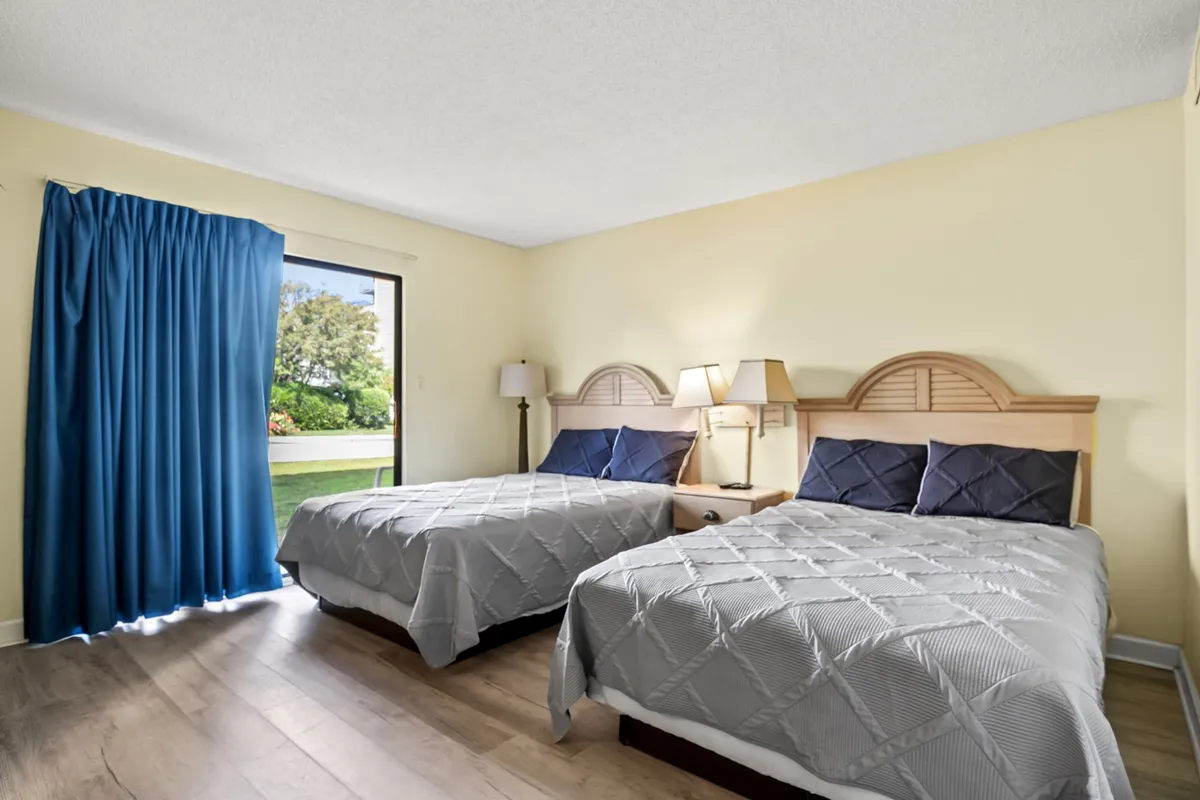 This scene shows two beds positioned near a large window with blue curtains, a nightstand nestled between them topped with lamps, and natural light streaming in, highlighting the sense of morning calm and rejuvenation.